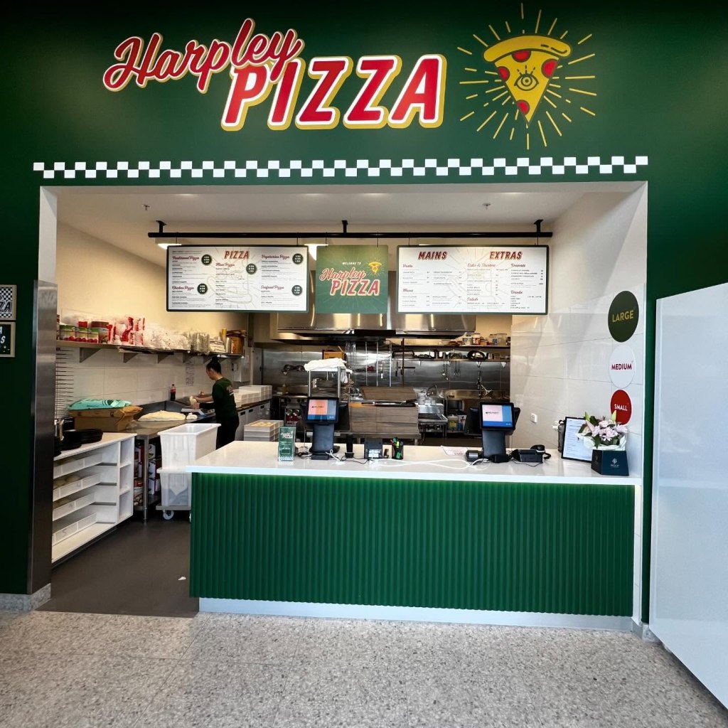 Pizza restaurant counter with green and white decor, menu above, and a staff member in the background. Signs for "Harpley Pizza" on the wall.